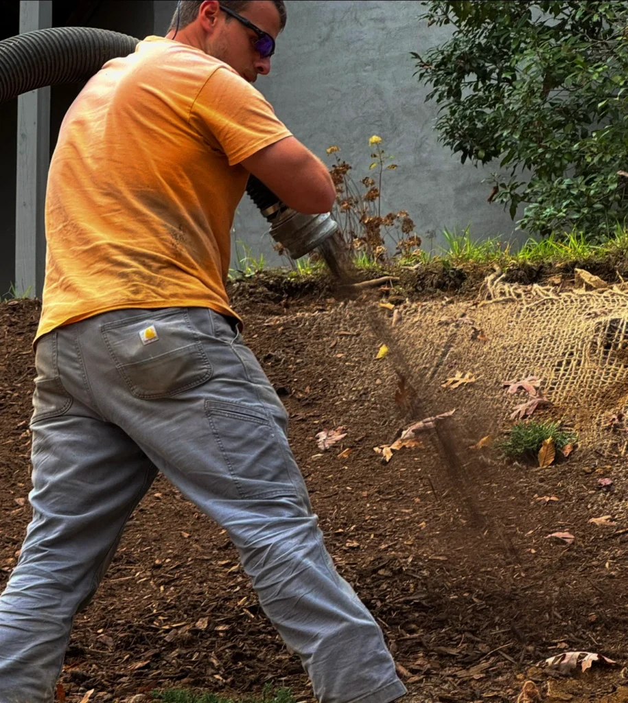 Close-up of a MulchWorx technician using a high-powered pneumatic hose to perform a blown mulch installation on a steep hillside embankment over erosion control matting.